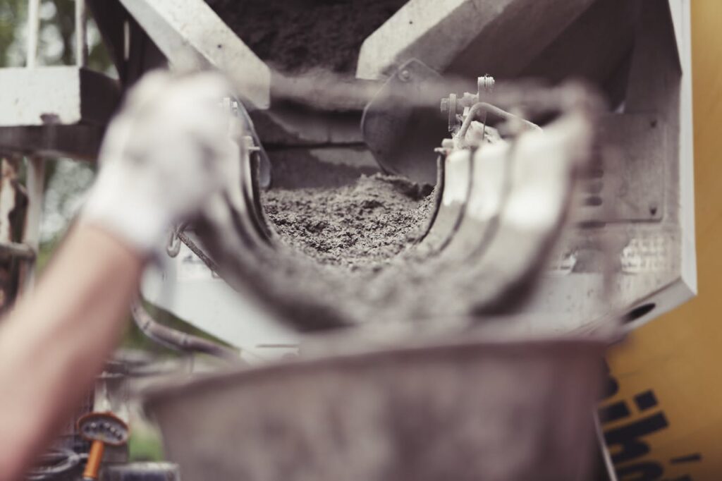 This picture shows the scene of a construction site. In the foreground a hand wearing work gloves is carrying a bucket. In the background a cement truck is visible. Cement is flowing from the truck into the bucket.